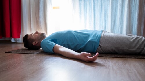 Young man meditating on a floor and lying in Shavasana pose at his living room.
