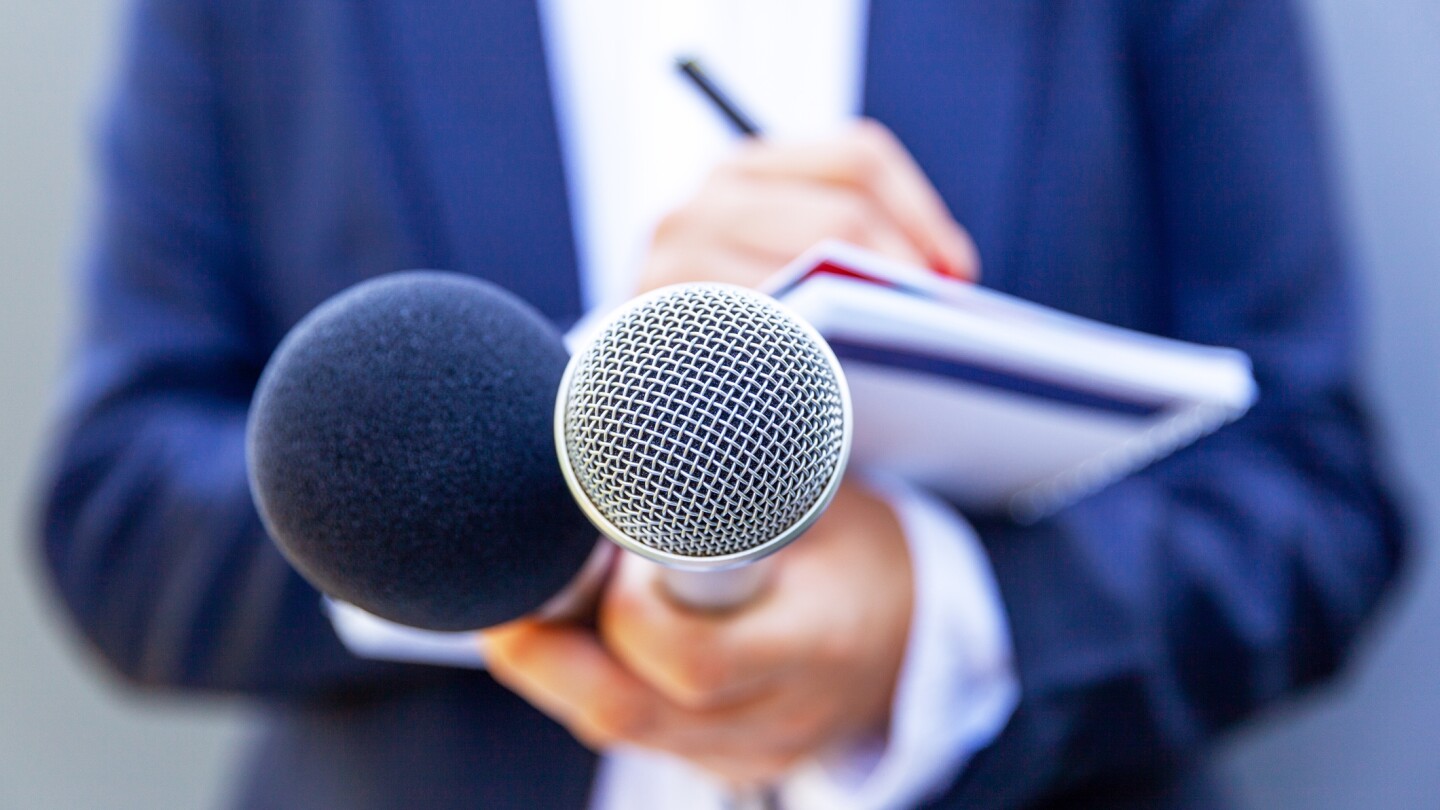 Microphone in the focus, news reporter at media event or press conference, holding mic, writing notes. Broadcast journalism concept.
