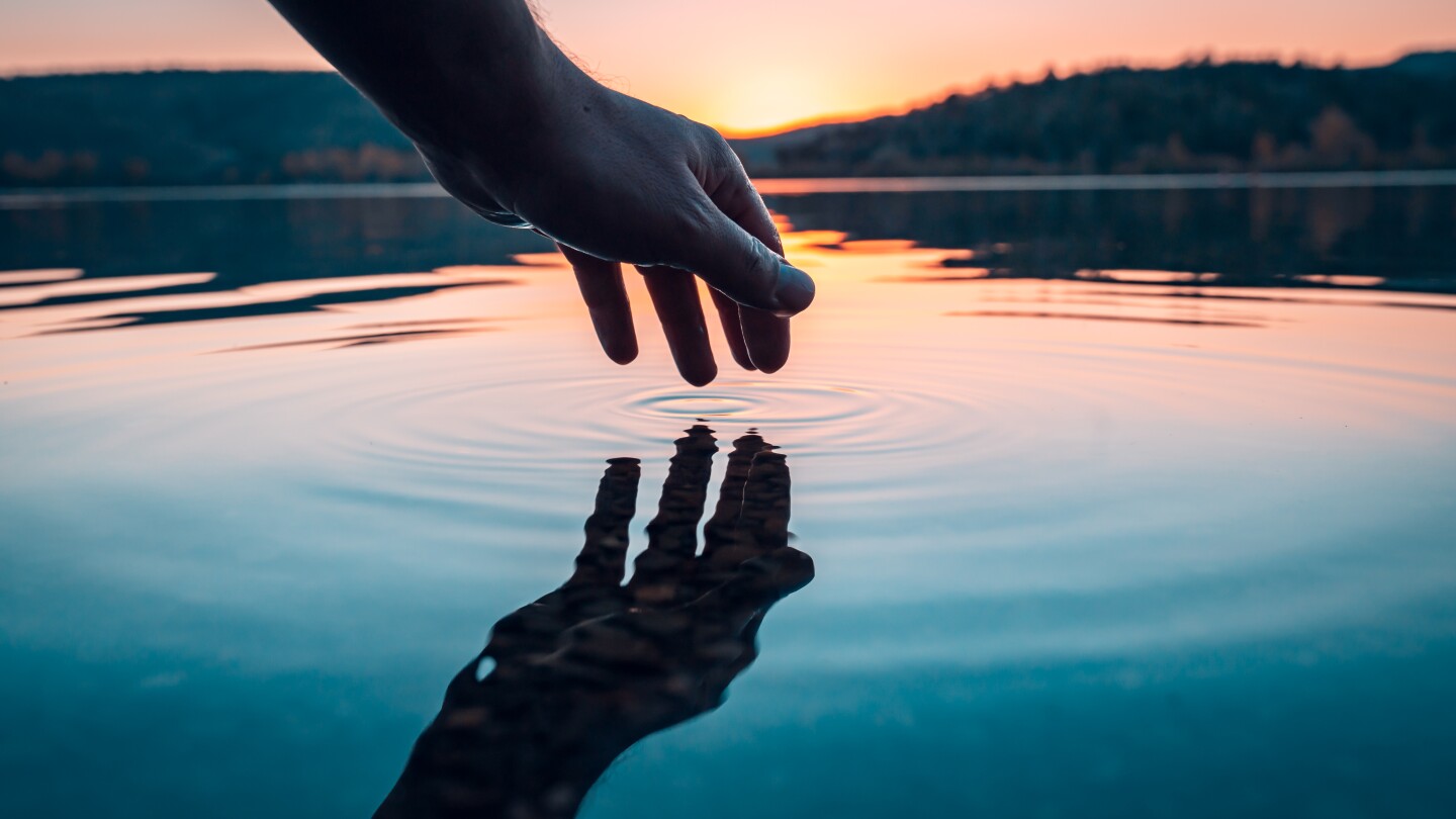 Finger touches surface of mountain lake. Hand reflection