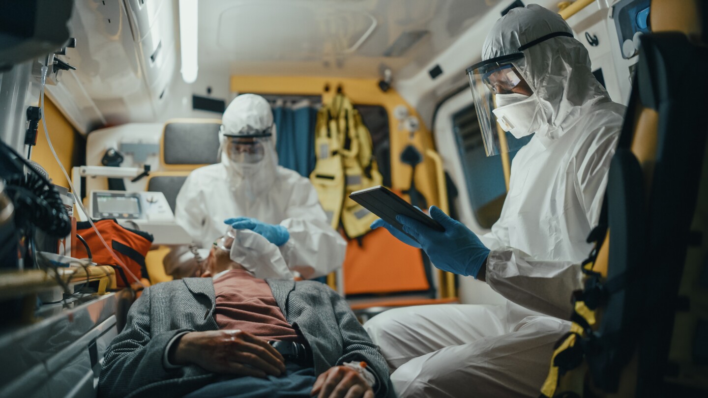 Paramedic in Coveralls Using Tablet Computer to Check the Diagnosis for the Injured Patient on the Way to Hospital. Emergency Medical Care Assistants Puts a Ventilation Mask in an Ambulance.