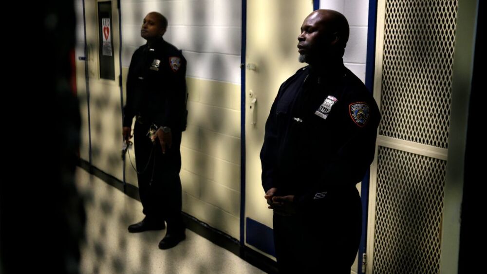 Two male correctional officers are positioned in a hallway, appearing attentive and ready for duty.
